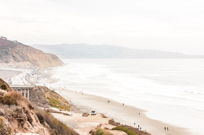 A slightly hazy, high-angle Landscape Beach Print captures a wide beachscape print where a light blue ocean waves meet a sandy shore dotted with numerous people enjoying the Summer Vibes. Gentle white waves roll towards the beach. Blue Lifeguard Towers line the coast in the far distance, of this framed landscape beach print. The overall lighting suggests a bright, overcast travel photography day on the San Diego coast. 