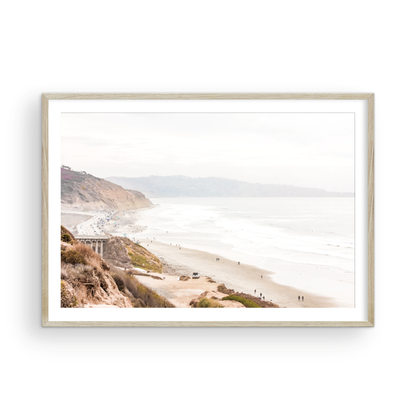 A slightly hazy, high-angle Landscape Beach Print captures a wide beachscape print where a light blue ocean waves meet a sandy shore dotted with numerous people enjoying the Summer Vibes. Gentle white waves roll towards the beach. Blue Lifeguard Towers line the coast in the far distance, of this framed landscape beach print. The overall lighting suggests a bright, overcast travel photography day on the San Diego coast. Photograph in minimalist mock up