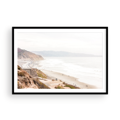 A slightly hazy, high-angle Landscape Beach Print captures a wide beachscape print where a light blue ocean waves meet a sandy shore dotted with numerous people enjoying the Summer Vibes. Gentle white waves roll towards the beach. Blue Lifeguard Towers line the coast in the far distance, of this framed landscape beach print. The overall lighting suggests a bright, overcast travel photography day on the San Diego coast. Photograph in minimalist mock up
