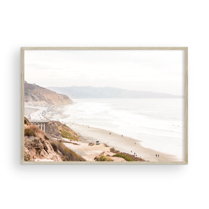 A slightly hazy, high-angle Landscape Beach Print captures a wide beachscape print where a light blue ocean waves meet a sandy shore dotted with numerous people enjoying the Summer Vibes. Gentle white waves roll towards the beach. Blue Lifeguard Towers line the coast in the far distance, of this framed landscape beach print. The overall lighting suggests a bright, overcast travel photography day on the San Diego coast. Photograph in minimalist mock up