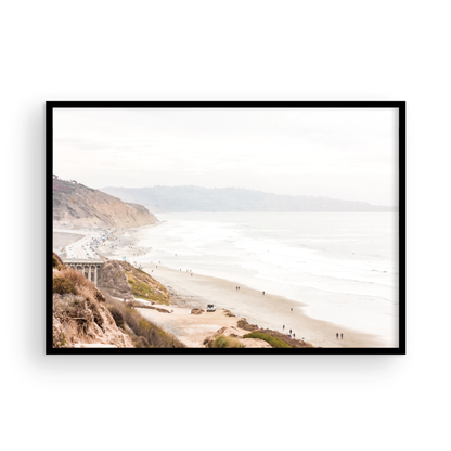 A slightly hazy, high-angle Landscape Beach Print captures a wide beachscape print where a light blue ocean waves meet a sandy shore dotted with numerous people enjoying the Summer Vibes. Gentle white waves roll towards the beach. Blue Lifeguard Towers line the coast in the far distance, of this framed landscape beach print. The overall lighting suggests a bright, overcast travel photography day on the San Diego coast. Photograph in minimalist mock up