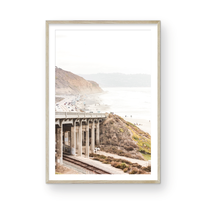 Captivating Torrey Pines San Diego coast scene, presented as vertical beach art. This piece of travel photography features a prominent bridge extending over a wide sandy beach with calm ocean waves. A coastal road winds along the base of rugged cliffs, with a lone coastal palm tree adding a touch of tropical essence to this Southern California seascape print. Photograph in minimalist mock up