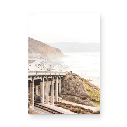 Captivating Torrey Pines San Diego coast scene, presented as vertical beach art. This piece of travel photography features a prominent bridge extending over a wide sandy beach with calm ocean waves. A coastal road winds along the base of rugged cliffs, with a lone coastal palm tree adding a touch of tropical essence to this Southern California seascape print. Photograph in minimalist unframed mock up