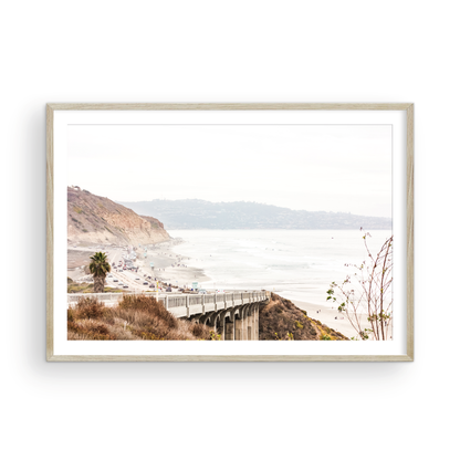 Captivating San Diego coast scene, presented as horizontal beach art. This piece of travel photography features a prominent bridge extending over a wide sandy beach with calm ocean waves. A coastal road winds along the base of rugged cliffs, with a lone coastal palm tree adding a touch of tropical essence to this Southern California seascape print. Photograph in minimalist mock up