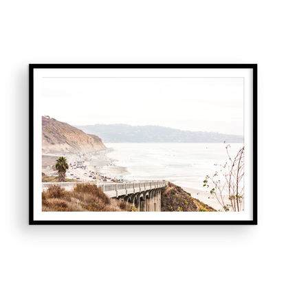 Captivating San Diego coast scene, presented as horizontal beach art. This piece of travel photography features a prominent bridge extending over a wide sandy beach with calm ocean waves. A coastal road winds along the base of rugged cliffs, with a lone coastal palm tree adding a touch of tropical essence to this Southern California seascape print. Photograph in minimalist mock up