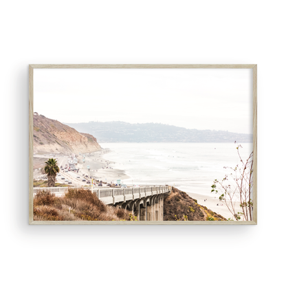 Captivating San Diego coast scene, presented as horizontal beach art. This piece of travel photography features a prominent bridge extending over a wide sandy beach with calm ocean waves. A coastal road winds along the base of rugged cliffs, with a lone coastal palm tree adding a touch of tropical essence to this Southern California seascape print. Photograph in minimalist mock up