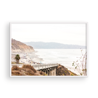Captivating San Diego coast scene, presented as horizontal beach art. This piece of travel photography features a prominent bridge extending over a wide sandy beach with calm ocean waves. A coastal road winds along the base of rugged cliffs, with a lone coastal palm tree adding a touch of tropical essence to this Southern California seascape print. Photograph in minimalist mock up