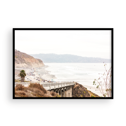 Captivating San Diego coast scene, presented as horizontal beach art. This piece of travel photography features a prominent bridge extending over a wide sandy beach with calm ocean waves. A coastal road winds along the base of rugged cliffs, with a lone coastal palm tree adding a touch of tropical essence to this Southern California seascape print. Photograph in minimalist mock up