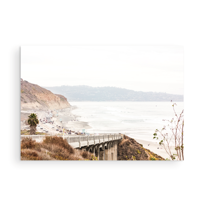 Captivating San Diego coast scene, presented as horizontal beach art. This piece of travel photography features a prominent bridge extending over a wide sandy beach with calm ocean waves. A coastal road winds along the base of rugged cliffs, with a lone coastal palm tree adding a touch of tropical essence to this Southern California seascape print. Photograph in minimalist mock up