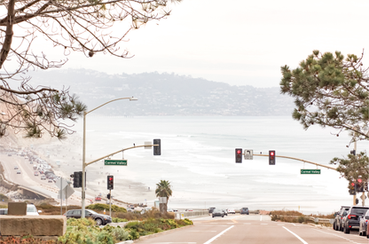 A hazy eye-level shot of San Diego Streets looks down a sloping road featuring red traffic lights and "Carmel Valley" signs, capturing a Local Surf Print vibe. The foreground of this California Wall Art showcases landscape and cars, framed by tree branches at the top, adding to the San Diego Photo aesthetic. The lighting suggests a hazy daytime, perfect for Minimalist Beach Wall Art. This horizontal San Diego print makes a great piece of horizontal beach wall decor. 