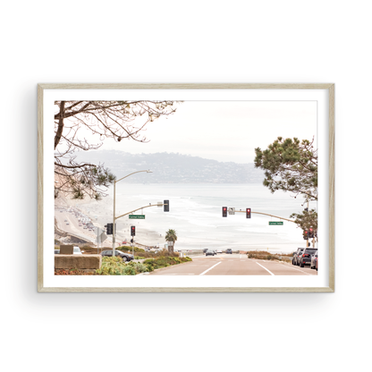 A hazy eye-level shot of San Diego Streets looks down a sloping road featuring red traffic lights and "Carmel Valley" signs, capturing a Local Surf Print vibe. The foreground of this California Wall Art showcases landscape and cars, framed by tree branches at the top, adding to the San Diego Photo aesthetic. The lighting suggests a daytime, perfect for Minimalist Beach Wall Art. This horizontal San Diego print makes a great piece of horizontal beach wall decor. Photograph in minimalist mock up