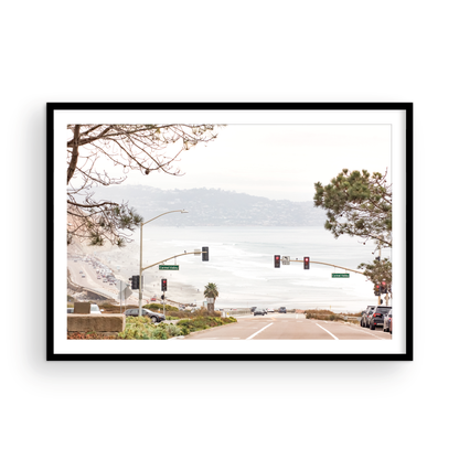 A hazy eye-level shot of San Diego Streets looks down a sloping road featuring red traffic lights and "Carmel Valley" signs, capturing a Local Surf Print vibe. The foreground of this California Wall Art showcases landscape and cars, framed by tree branches at the top, adding to the San Diego Photo aesthetic. The lighting suggests a daytime, perfect for Minimalist Beach Wall Art. This horizontal San Diego print makes a great piece of horizontal beach wall decor. Photograph in minimalist mock up