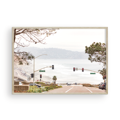 A hazy eye-level shot of San Diego Streets looks down a sloping road featuring red traffic lights and "Carmel Valley" signs, capturing a Local Surf Print vibe. The foreground of this California Wall Art showcases landscape and cars, framed by tree branches at the top, adding to the San Diego Photo aesthetic. The lighting suggests a daytime, perfect for Minimalist Beach Wall Art. This horizontal San Diego print makes a great piece of horizontal beach wall decor. Photograph in minimalist mock up