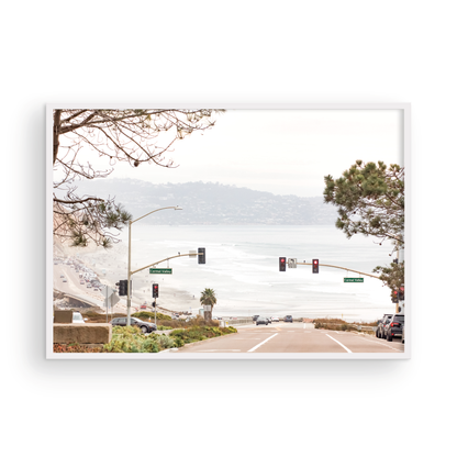 A hazy eye-level shot of San Diego Streets looks down a sloping road featuring red traffic lights and "Carmel Valley" signs, capturing a Local Surf Print vibe. The foreground of this California Wall Art showcases landscape and cars, framed by tree branches at the top, adding to the San Diego Photo aesthetic. The lighting suggests a daytime, perfect for Minimalist Beach Wall Art. This horizontal San Diego print makes a great piece of horizontal beach wall decor. Photograph in minimalist mock up