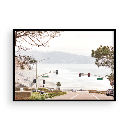 A hazy eye-level shot of San Diego Streets looks down a sloping road featuring red traffic lights and "Carmel Valley" signs, capturing a Local Surf Print vibe. The foreground of this California Wall Art showcases landscape and cars, framed by tree branches at the top, adding to the San Diego Photo aesthetic. The lighting suggests a daytime, perfect for Minimalist Beach Wall Art. This horizontal San Diego print makes a great piece of horizontal beach wall decor. Photograph in minimalist mock up