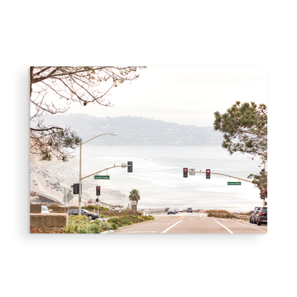 A hazy eye-level shot of San Diego Streets looks down a sloping road featuring red traffic lights and "Carmel Valley" signs, capturing a Local Surf Print vibe. The foreground of this California Wall Art showcases landscape and cars, framed by tree branches at the top, adding to the San Diego Photo aesthetic. The lighting suggests a daytime, perfect for Minimalist Beach Wall Art. This horizontal San Diego print makes a great piece of horizontal beach wall decor. Photograph in minimalist mock up