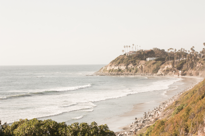 Fine art of Swamis Beach, San Diego California horizontal print of hazy high-angle landscape beach photography captures a light blue ocean meeting a sandy beach with many beach goers. To the right, tall tan and brown cliffs rise as iconic coastal scenery. 