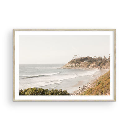 Fine art of Swamis Beach, San Diego California horizontal print of hazy high-angle landscape beach photography captures a light blue ocean meeting a sandy beach with many beach goers. To the right, tall tan and brown cliffs rise as iconic coastal scenery. Photograph in minimalist framed mock up.