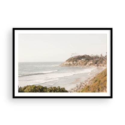 Fine art of Swamis Beach, San Diego California horizontal print of hazy high-angle landscape beach photography captures a light blue ocean meeting a sandy beach with many beach goers. To the right, tall tan and brown cliffs rise as iconic coastal scenery. Photograph in minimalist framed mock up.