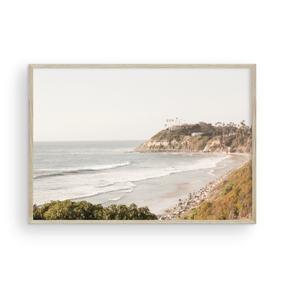 Fine art of Swamis Beach, San Diego California horizontal print of hazy high-angle landscape beach photography captures a light blue ocean meeting a sandy beach with many beach goers. To the right, tall tan and brown cliffs rise as iconic coastal scenery. Photograph in minimalist framed mock up.