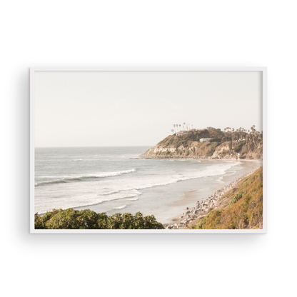 Fine art of Swamis Beach, San Diego California horizontal print of hazy high-angle landscape beach photography captures a light blue ocean meeting a sandy beach with many beach goers. To the right, tall tan and brown cliffs rise as iconic coastal scenery. Photograph in minimalist framed mock up.