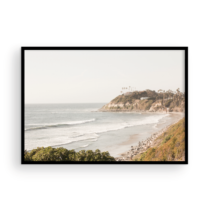 Fine art of Swamis Beach, San Diego California horizontal print of hazy high-angle landscape beach photography captures a light blue ocean meeting a sandy beach with many beach goers. To the right, tall tan and brown cliffs rise as iconic coastal scenery. Photograph in minimalist framed mock up.