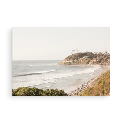 Fine art of Swamis Beach, San Diego California horizontal print of hazy high-angle landscape beach photography captures a light blue ocean meeting a sandy beach with many beach goers. To the right, tall tan and brown cliffs rise as iconic coastal scenery. Photograph in minimalist framed mock up.