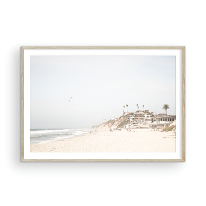 A bright, minimalist photograph of Moonlight State Beach in Encinitas, California, showing a wide sandy beach, gentle ocean waves, and residential buildings atop a cliff on the right, with a seagull flying in the hazy sky. Photograph in minimalist mock up