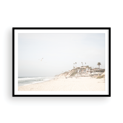 A bright, minimalist photograph of Moonlight State Beach in Encinitas, California, showing a wide sandy beach, gentle ocean waves, and residential buildings atop a cliff on the right, with a seagull flying in the hazy sky. Photograph in minimalist mock up