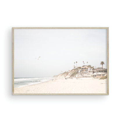 A bright, minimalist photograph of Moonlight State Beach in Encinitas, California, showing a wide sandy beach, gentle ocean waves, and residential buildings atop a cliff on the right, with a seagull flying in the hazy sky. Photograph in minimalist mock up