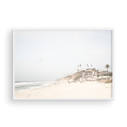 A bright, minimalist photograph of Moonlight State Beach in Encinitas, California, showing a wide sandy beach, gentle ocean waves, and residential buildings atop a cliff on the right, with a seagull flying in the hazy sky. Photograph in minimalist mock up