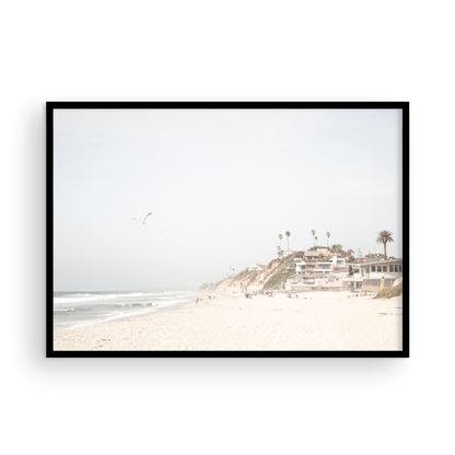 A bright, minimalist photograph of Moonlight State Beach in Encinitas, California, showing a wide sandy beach, gentle ocean waves, and residential buildings atop a cliff on the right, with a seagull flying in the hazy sky. Photograph in minimalist mock up