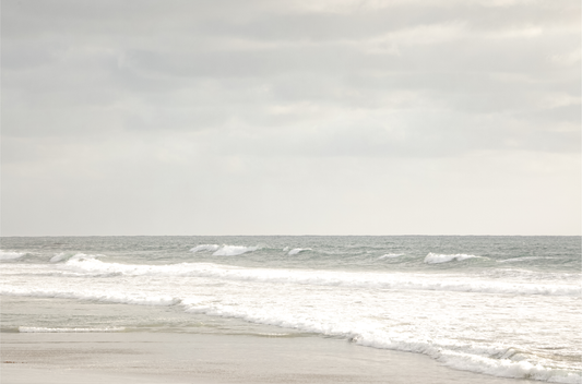 Horizontal minimalist ocean print of gentle ocean waves rolling onto a sandy shoreline at Moonlight Beach, San Diego, under a bright, soft sky. This calm beach print provides a serene seascape. 