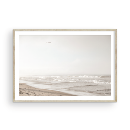 Minimalist ocean print featuring gentle ocean waves breaking on a sandy beach under a bright, hazy sky with a single bird in flight. A serene and neutral ocean print. Photograph in minimalist mock up.