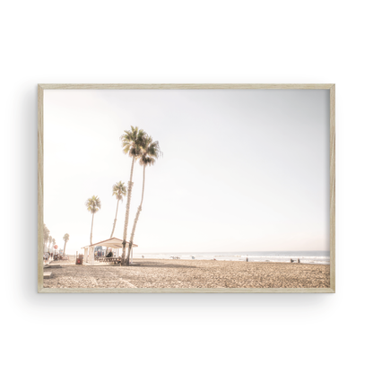 Landscape beach photography of Venice Beach, California, showcasing the sandy beach, ocean, and a small shelter with palm trees along the boardwalk. Captures the casual beach art feel. Photograph in minimalist mock up.