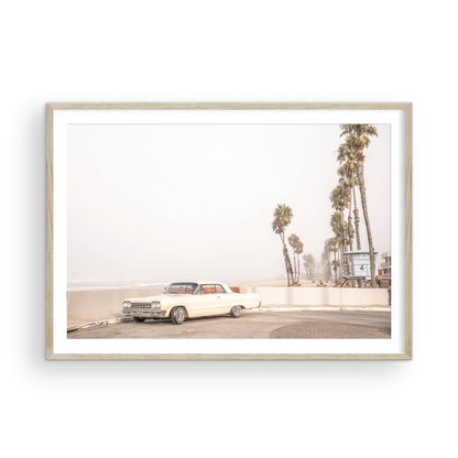 Landscape beach photography featuring a vintage white car parked on a coastal road with tall palm trees and a lifeguard tower nearby, overlooking the ocean. Photograph in minimalist mock up.