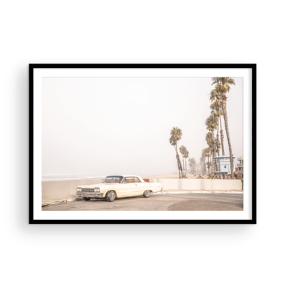 Landscape beach photography featuring a vintage white car parked on a coastal road with tall palm trees and a lifeguard tower nearby, overlooking the ocean. Photograph in minimalist mock up.