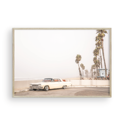 Landscape beach photography featuring a vintage white car parked on a coastal road with tall palm trees and a lifeguard tower nearby, overlooking the ocean. Photograph in minimalist mock up.