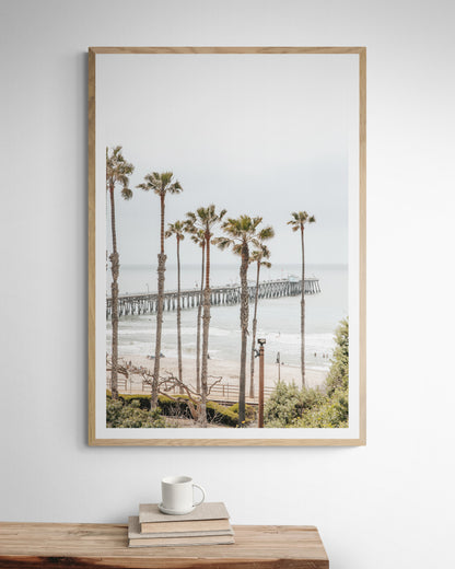 A vertical landscape coastal photography shot of the San Clemente Pier from behind a lush line of tall palm trees. This beachscape print highlights the California coast and is ideal for large beach art or a beach theme wall decor, with subtle ocean waves. Photograph in minimalist mock up.
