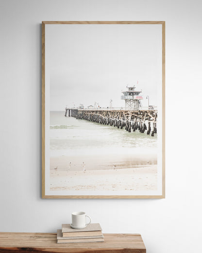 A long wooden pier, the San Clemente Pier, extends into the ocean in this vertical coastal photography. The beach in the foreground suggests a calm ocean print, ideal for a large coastal wall art or a beach cottage wall decor. Photograph in minimalist mock up.