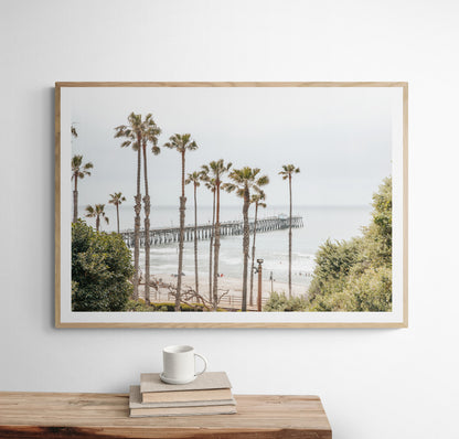 A horizontal landscape coastal photography shot of the San Clemente Pier from behind a lush line of tall palm trees. This beachscape print highlights the California coast and is ideal for large landscape beach art or a beach theme wall decor, with subtle ocean waves. Photograph in minimalist mock up.
