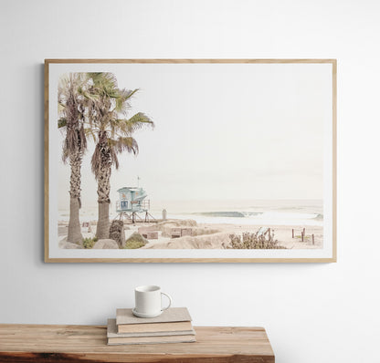 Dreamy coastal palm tree print featuring two tall palm trees with shaggy trunks in the foreground and a pastel blue lifeguard tower in the distance. This horizontal tropical print is captured at Seaside Beach on the Southern California coast, is bathed in bright, hazy sunlight, creating a pastel beach art effect. Evokes serene summer vibes, perfect for beach house decor. Photograph in minimalist mock up.