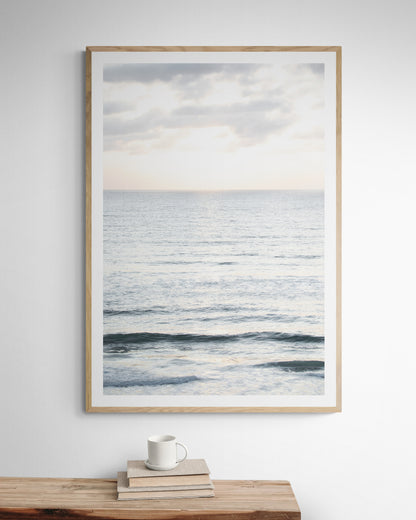 Fine art of vertical landscape print by Southern California photographer Kim Dybczak of Salt Creek Prints, featuring a bright image capturing pale blue ocean waves, light gray clouds in a bright sky. Gentle, small waves in the near and far distance. The sky is a uniform, pale white, creating a soft, overcast atmosphere. Photograph in framed minimalist mock up