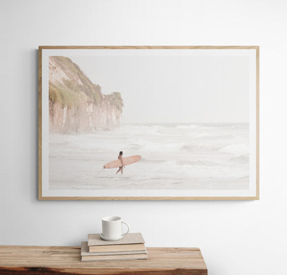 Horizontal surf art print of a lone surfer carrying a surfboard, walking on a rocky, foggy shoreline at Moonlight State Beach with a cliff in the background. Photograph in minimalist mock up.