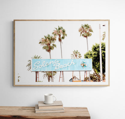 Colorful travel photography capturing the iconic 'Solana Beach' sign, a quintessential piece of Surf Signs San Diego art. The retro light blue sign, adorned with white cursive script and a unique steer head logo, is set against a backdrop of towering palm trees and a bright, hazy California sky. This Southern California surf beach sign evokes strong summer vibes and is perfect for coastal decor. Photograph in minimalist framed mock up.