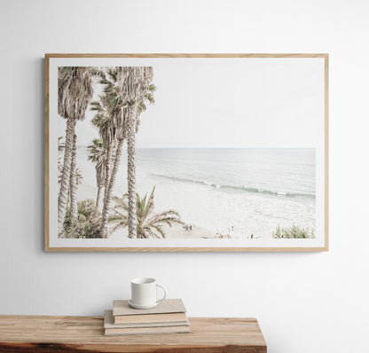 Dreamy coastal palm tree print featuring several tall palm trees with shaggy trunks, and surfers walking down the tranquil beach. This vertical tropical print is captured at Swamis Beach on the Southern California coast, is bathed in bright, hazy sunlight, creating a pastel beach art effect. Evokes serene summer vibes, perfect for beach house decor. Photograph in minimalist mock up.