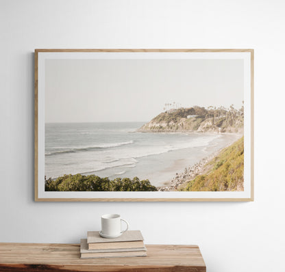 Fine art of Swamis Beach, San Diego California horizontal print of hazy high-angle landscape beach photography captures a light blue ocean meeting a sandy beach with many beach goers. To the right, tall tan and brown cliffs rise as iconic coastal scenery. Photograph in minimalist framed mock up.