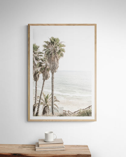 Dreamy coastal palm tree print featuring several tall palm trees with shaggy trunks, next to a staircase down to a tranquil beach. This vertical tropical print is captured at Swamis Beach on the Southern California coast, is bathed in bright, hazy sunlight, creating a pastel beach art effect. Evokes serene summer vibes, perfect for beach house decor. Photograph in minimalist surf art mock up.