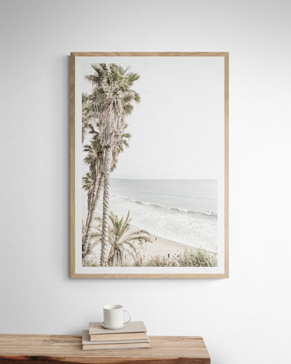 Dreamy coastal palm tree print featuring several tall palm trees with shaggy trunks, lining a gentle slope down to a tranquil beach. This vertical tropical print is captured at Swamis Beach on the Southern California coast, is bathed in bright, hazy sunlight, creating a pastel beach art effect. Evokes serene summer vibes, perfect for beach house decor. Photograph in minimalist surf art mock up.