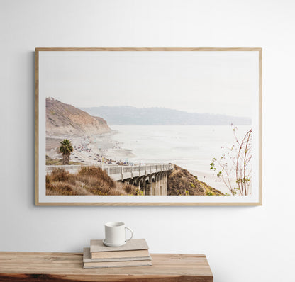 Captivating San Diego coast scene, presented as horizontal beach art. This piece of travel photography features a prominent bridge extending over a wide sandy beach with calm ocean waves. A coastal road winds along the base of rugged cliffs, with a lone coastal palm tree adding a touch of tropical essence to this Southern California seascape print. Photograph in minimalist mock up